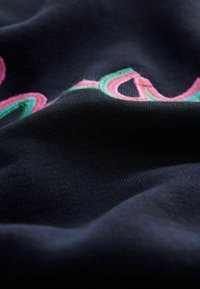 Tissu noir avec des lettres colorées brodées en relief en rose, vert et bleu, montrant la texture et des plis courbés.