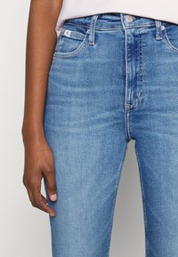 Persona che indossa jeans di denim azzurro chiaro con tasche frontali visibili e bottone in metallo, mano rilassata accanto alla coscia su uno sfondo neutro.