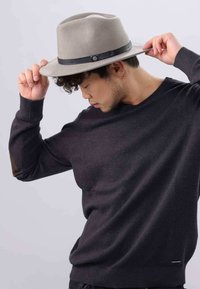 Un jeune homme ajustant un chapeau fedora beige, portant un pull sombre texturé avec des coudières, les yeux fermés et le regard dirigé vers le bas.