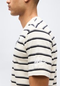 Πλαϊνή όψη ατόμου που φοράει μπεζ και μαύρο ριγέ κοντομάνικο πουκάμισο Nike με κεντημένο λογότυπο στο μανίκι, μπροστά σε απλή φόντο.