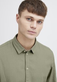 Lys grønne skjorte med knapper og krage. Tekstureret stof og synlige knapper. Fokus på skjortens front og pasform omkring halsen.