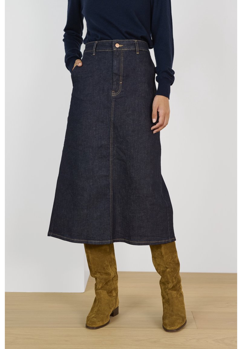 Femme portant une jupe midi en denim foncé avec des poches avant et des bottes hautes en daim camel, debout sur un sol en bois.