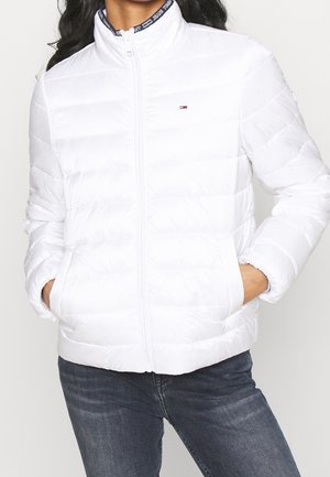 Kvinde iført en hvid quiltet jakke med frontlynlås og lommer, kombineret med mørkeblå jeans.