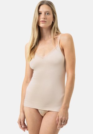 Kvinde med langt blondt hår iført en beige camisole og matchende undertøj, stående mod en ensartet lys baggrund.
