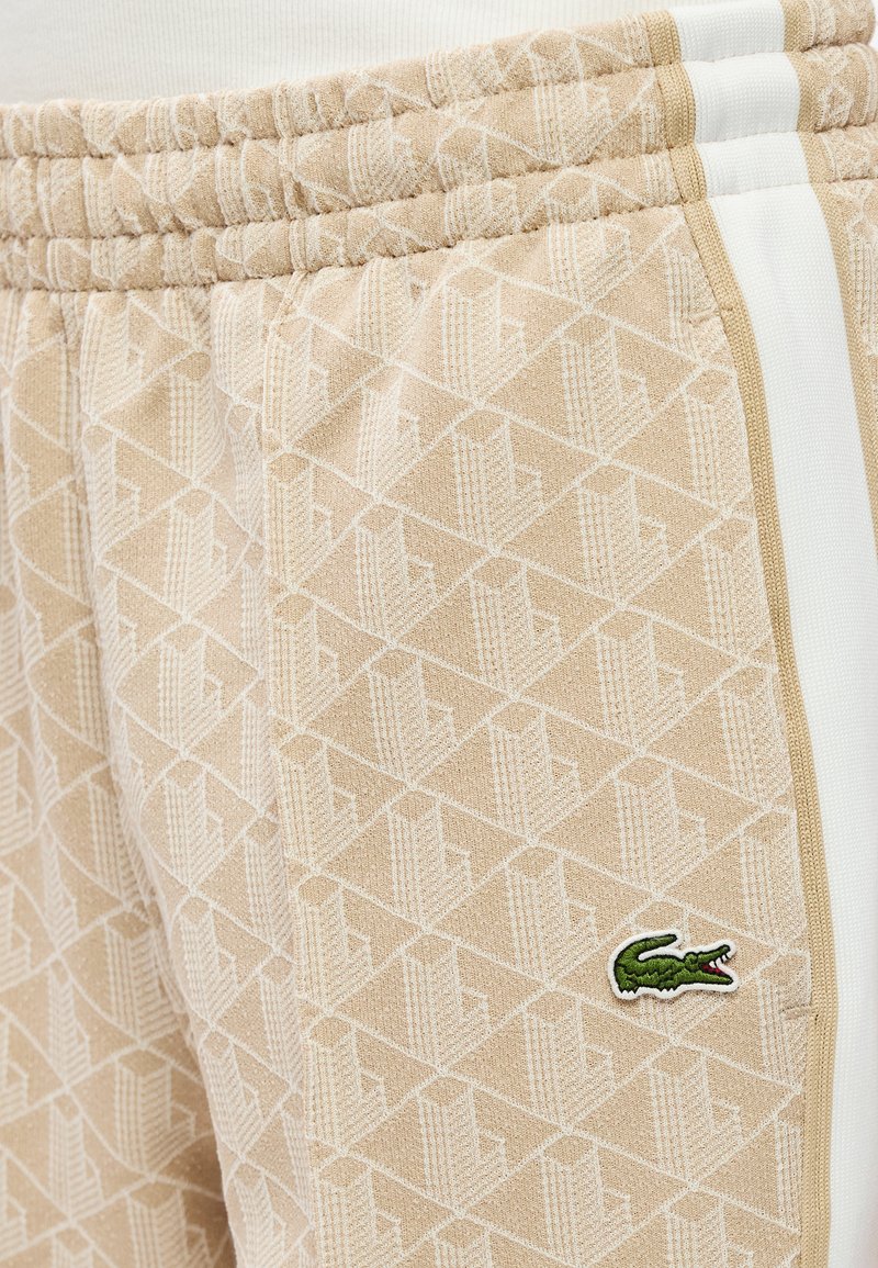 Pantaloni della tuta beige con motivo geometrico strutturato, caratterizzati da strisce bianche e un logo coccodrillo verde ricamato sul lato.