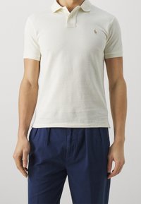 Man in een crèmekleurig polo shirt met korte mouwen en marineblauwe geplooide broek, staand tegen een effen lichte achtergrond.