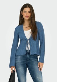 Hellblauer gerippter Cardigan mit einem Bindegürtel, langen Ärmeln und einem ausgestellten Saum, kombiniert mit einem weißen Oberteil und dunkelblauen Denim-Jeans.