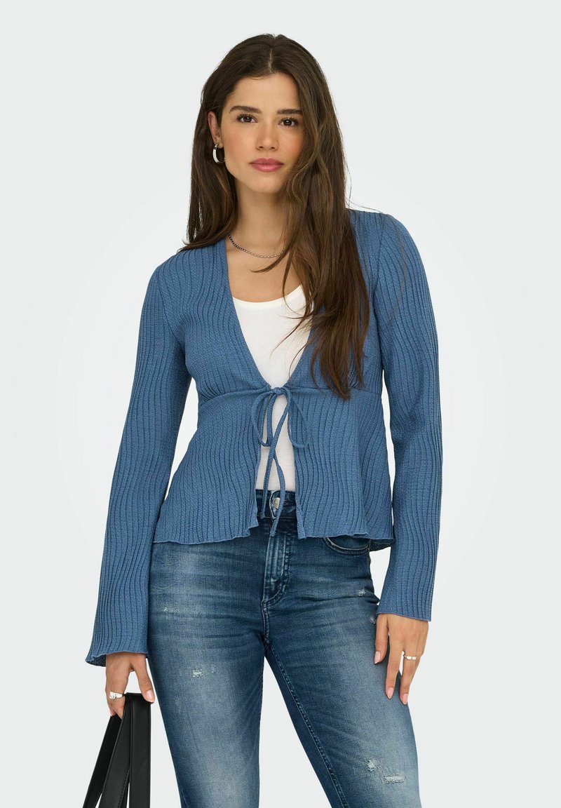 Hellblauer gerippter Cardigan mit einem Bindegürtel, langen Ärmeln und einem ausgestellten Saum, kombiniert mit einem weißen Oberteil und dunkelblauen Denim-Jeans.