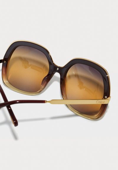 Gafas de sol con degradado marrón, con grandes monturas cuadradas y acentos dorados. Diseño de carey en los bordes, con un logo dorado en la patilla.