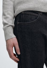 Jeans in denim scuro con una texture sottile, caratterizzati dallo stile classico a cinque tasche e cuciture gialle a contrasto. La mano poggia sulla coscia.