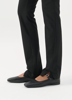 Piernas con pantalones ajustados negros con aberturas frontales en el dobladillo, combinados con zapatos planos Mary Jane negros con una pequeña correa con hebilla.