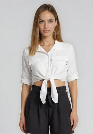 Jeune femme aux cheveux raides portant un crop top blanc noué et un short noir taille haute, debout devant un fond uni.