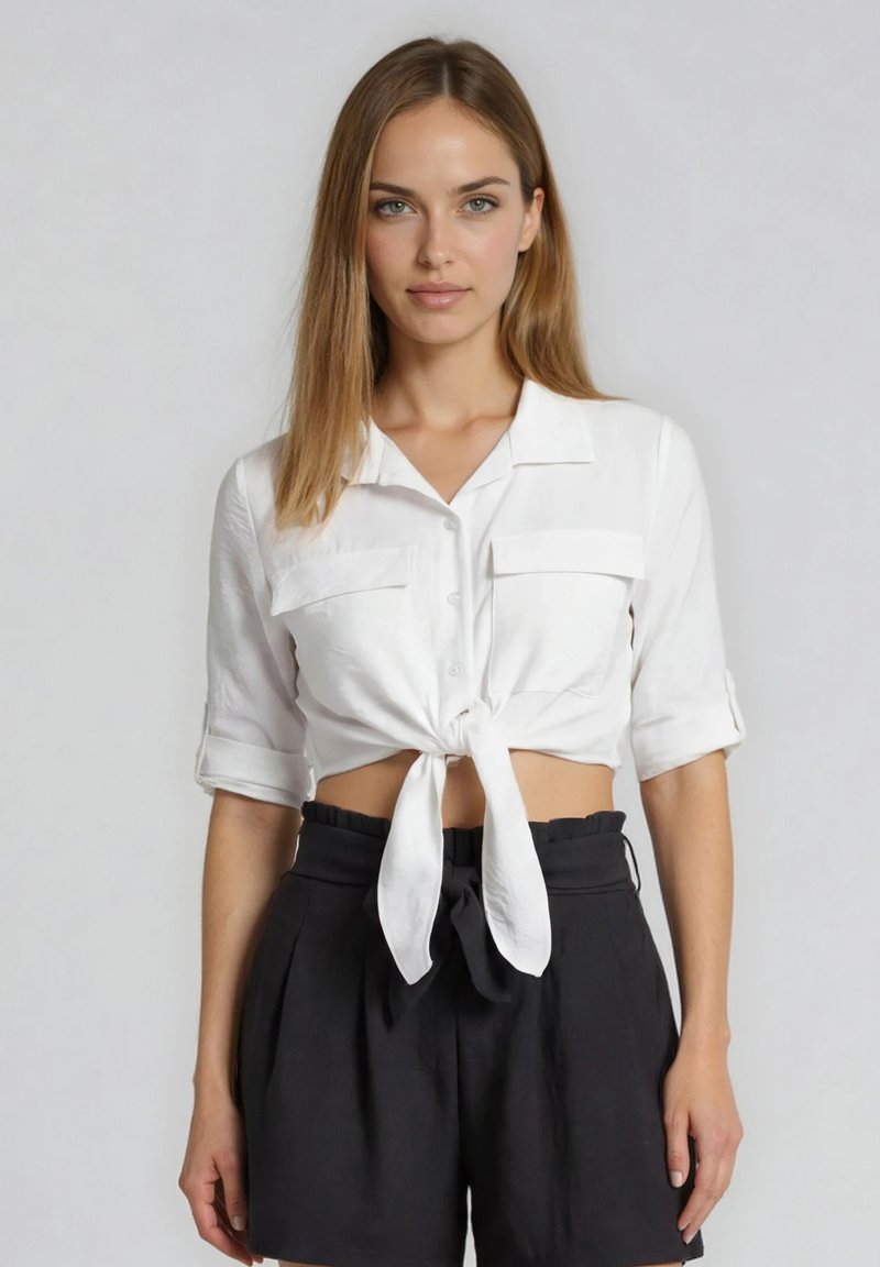 Νέα γυναίκα με ίσια μαλλιά που φοράει λευκό crop top με δέσιμο και μαύρα σορτς με ψηλή μέση, στέκεται μπροστά σε ένα απλό φόντο.