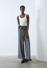 Pantalon large à rayures grises avec une taille nouée, associé à un débardeur blanc et des chaussures noires, sur fond neutre.