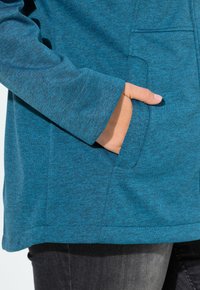 Henkilön käsi lepää sinisen pitkähihapaitojen etutaskussa, yhdistettynä tummanharmaisiin housuihin.