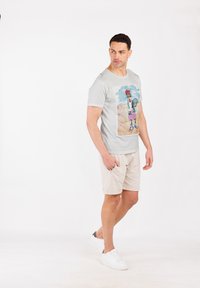 T-shirt grigio chiaro con una stampa grafica colorata di un personaggio, abbinato a pantaloni corti beige e sneaker bianche. Design semplice e informale.