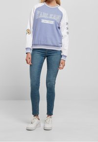 Světlomodrá mikina s bílými rukávy, zdobená textem „KARL KANI“ a nápisem založeno 1989. Má žebrované manžety a lem, kombinovaná s úzkými modrými džínami a bílými botami.