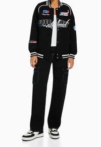 Zwarte varsityjack met witte strepen, kleurrijke patches en geborduurde tekst. Gecombineerd met zwarte cargo broeken en zwart-witte sneakers.