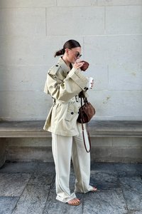 Beige lielgabarīta trencs ar jostu viduklī, krēmkrāsas plati bikses, brūna zamšādas rokassoma un saulesbrilles. Tur rokā kafiju un maizīti.