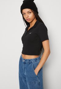 Črna polo majica s kratkimi rokavi in ovratnikom ter belim logotipom, v kombinaciji z modrimi kavbojkami, ki imajo žepe in elastičen pas.