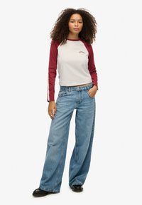 Witte en bordeaux lange mouwen shirt met ronde hals, gecombineerd met lichtblauwe wijde jeans en zwarte schoenen, tegen een witte achtergrond.