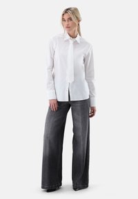 Camicia bianca a maniche lunghe con colletto appuntito e cravatta bianca, abbinata a pantaloni denim grigi a gamba larga con una texture sbiadita.