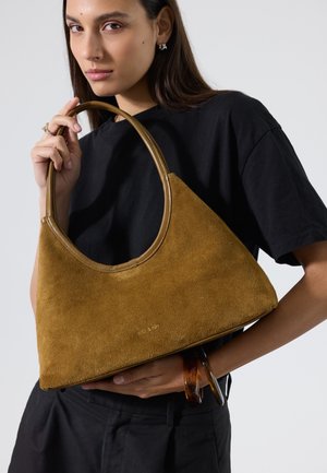Femme en chemise noire tenant un sac à main en daim marron avec des anses rondes, face à un fond clair uni.