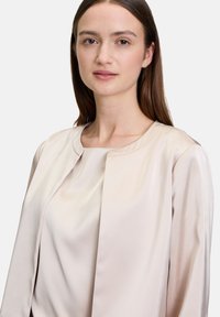 Beige Satinbluse mit rundem Ausschnitt, offenem Vorderteil und langen Ärmeln. Glatte Textur mit subtiler Ausstrahlung und minimalen Nähten.