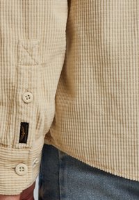Helle beige Corduroy-Hemden mit einer strukturierten Oberfläche, Knopfverschluss und einem schwarzen Etikett mit einem Logo am Saum.
