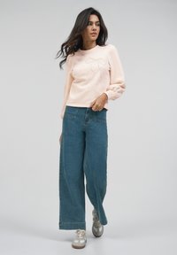 Lys rosa hettegenser med en sløyfedesign og dekorasjoner, sammen med brede blå jeans og grå joggesko. Glatt stoff og løs passform.