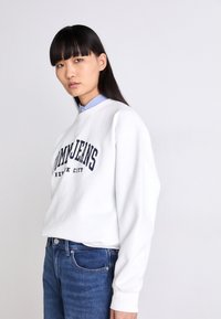 Sudadera blanca con "Tommy Jeans New York City" en letras azules marino, combinada con una camisa de cuello azul claro y jeans azules. Corte relajado, mangas largas.