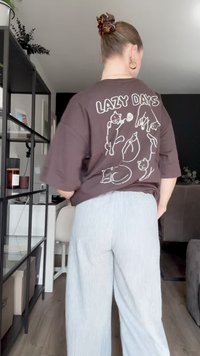 T-shirt oversized castanho com ilustrações de gatos brancos e o texto "LAZY DAYS" nas costas; combinado com calças largas de riscas claras.