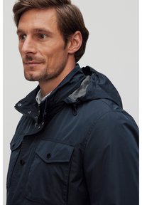 Laivastonsininen vedenpitävä takki, jossa on irrotettava huppu, kaksi etutaskua, nepparit ja sileä tekstiili, päällä kaulustakki.