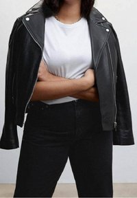 Personne portant une veste en cuir noire drapée sur les épaules, un t-shirt blanc et un pantalon noir, bras croisés devant un fond uni.