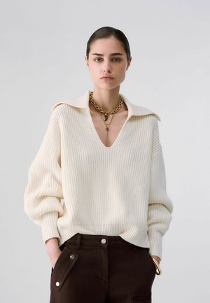 Femme portant un pull en maille crème avec un large col, un pantalon foncé avec des poches et des colliers en chaîne dorés superposés.