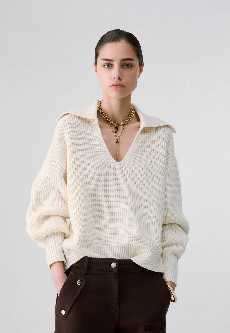 Femme portant un pull en maille crème avec un large col, un pantalon foncé avec des poches et des colliers en chaîne dorés superposés.