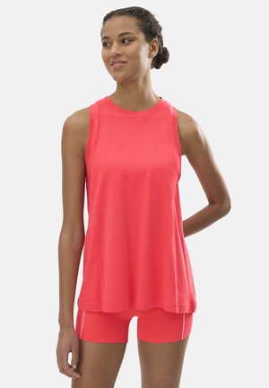 Mujer de pie con camiseta sin mangas roja y shorts ajustados a juego, mirando ligeramente hacia su izquierda con una suave sonrisa.