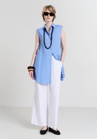 Persona che indossa una tunica senza maniche blu chiaro, pantaloni bianchi a gamba larga, ballerine nere, collana scura e braccialetti, in piedi su uno sfondo uniforme.