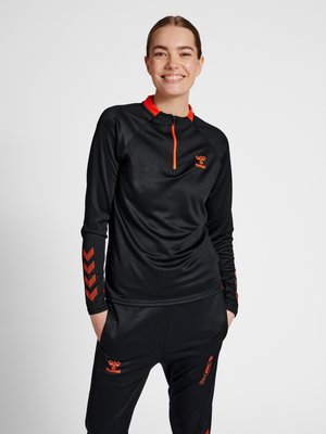 Donna sorridente che indossa abbigliamento sportivo nero e arancione con maniche lunghe e pantaloni, in piedi con le mani in tasca su uno sfondo semplice.