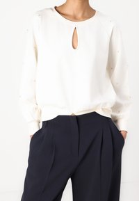 Persoon draagt een witte blouse met lange mouwen en een sleutelgat aan de voorkant en zwarte, hooggetailleerde, geplooide broek, handen in de zakken.