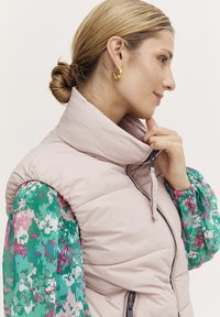 Vesta pufo roz deschis cu guler înalt, având un fermoar frontal și mâneci cu un model floral și de verdeață contrasting. Cercei de aur vizibili.