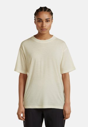 Ung person med flettet hår iført en simpel cremefarvet T-shirt med korte ærmer og sorte bukser, stående mod en hvid baggrund.