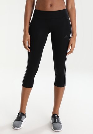 Person trägt schwarze, kurze Adidas-Leggings mit weißen Seitenstreifen und graue Slip-On-Sportschuhe und steht vor einem einfarbigen Hintergrund.