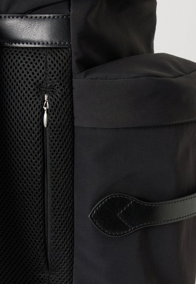 Gros plan sur un sac à dos noir montrant une poche en mesh avec une fermeture éclair et une poche latérale solide avec un détail de sangle en cuir.
