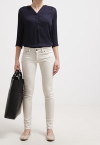 Marinblå knapprad blus med v-ringning, kombinerad med ljusfärgade åtsittande jeans och beige lägenheter, medan hon håller en svart tote-väska.