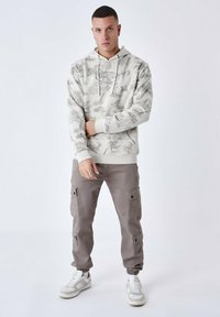 Felpa con cappuccio in beige chiaro con grafica grigia; abbinata a pantaloni cargo color taupe e sneaker bianche. Design semplice e casual.