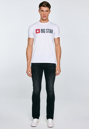 Młody mężczyzna stoi, ubrany w białą koszulkę "BIG STAR", czarne jeansy i białe trampki na jednolitym jasnym tle.