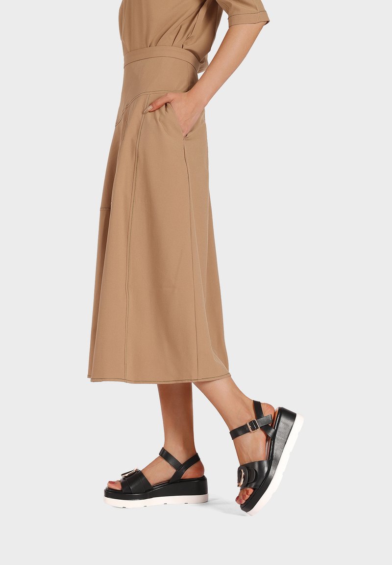 Abito midi color sabbia con vita cintrata e tasche, abbinato a sandali neri con plateau e dettagli con fibbia.