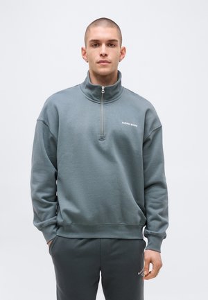 Fiatal férfi rövid hajjal, szürke, félig cipzáras pulóvert és hozzáillő nadrágot visel, mindkettőn kis fehér "Björn Borg" logók találhatók.