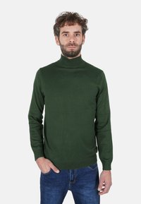 Maglione a collo alto verde scuro realizzato in morbido tessuto lavorato a maglia, con maniche lunghe e design aderente. Indossato con jeans blu.